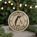 A round ornament with the name "Adelle Penguin" on it, featuring a penguin illustration, is displayed in front of a Christmas tree with twinkling lights.