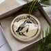 A decorative ornament featuring an illustration of an African penguin, placed in a box with pine branches.