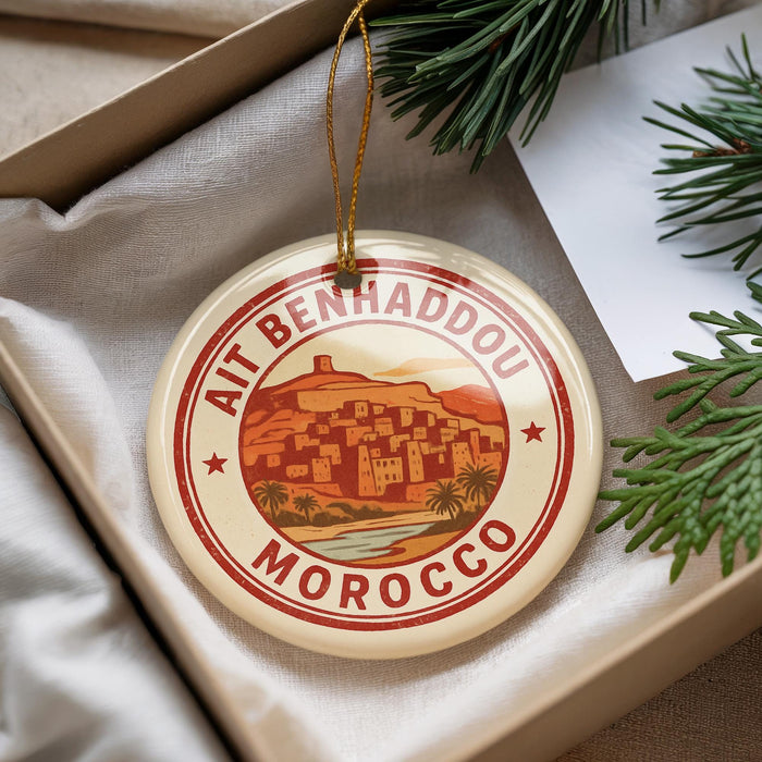 A circular ornament with a red and white design, featuring a desert town and mountains, is displayed in a box with pine branches.