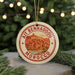 A circular ornament with a red border and a graphic of a city, surrounded by greenery and twinkling lights.