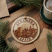 A circular ornament with the text "ALHAMBRA SPAIN" on it, featuring an illustration of a cityscape, is displayed on a wooden surface alongside a cup of coffee and some pine branches.