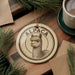 A circular ornament featuring an image of an alpaca is displayed on a wooden surface, accompanied by a cup of coffee and pine branches.