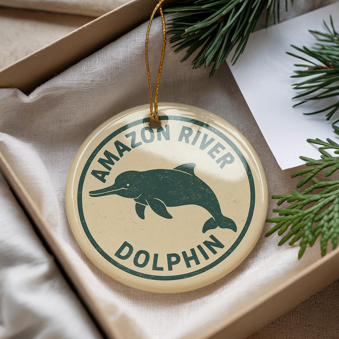 A round ornament with a green dolphin and the text "AMAZON RIVER DOLPHIN" is displayed in a box, surrounded by pine branches.