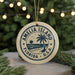 A circular ornament with a palm tree and beach scene, surrounded by a string of lights, placed on a surface with pine branches.