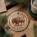 A circular ornament featuring an illustration of a bison, with the text "AMERICAN BISON" printed on it, is displayed on a wooden surface alongside pine branches and a cup of coffee.