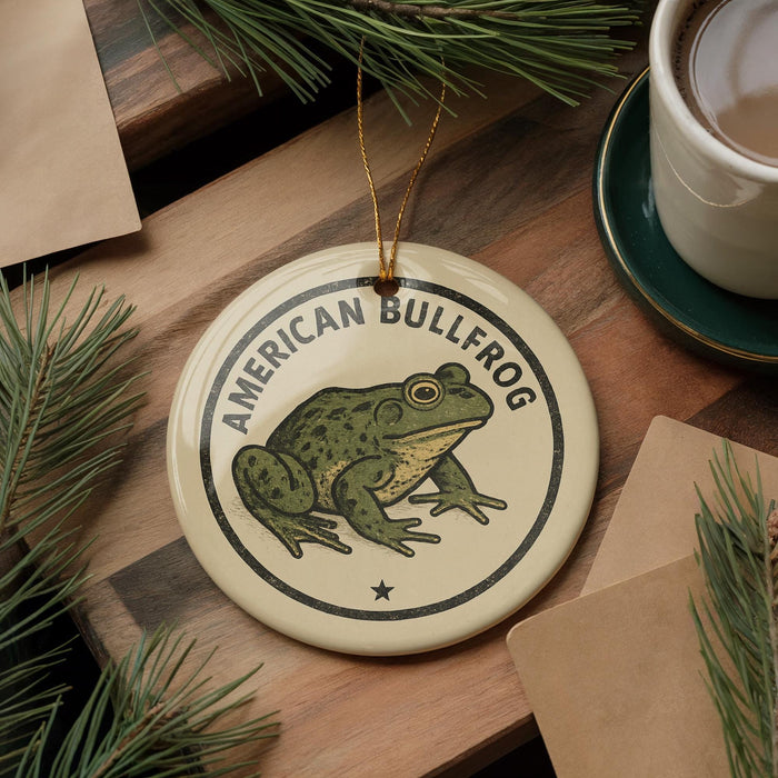 A circular ornament featuring an illustration of an American bullfrog, with the text "AMERICAN BULLFROG" printed on it, is displayed on a wooden surface alongside a cup of coffee and some pine branches.
