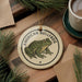 A circular ornament featuring an illustration of an American bullfrog, with the text "AMERICAN BULLFROG" printed on it, is displayed on a wooden surface alongside a cup of coffee and some pine branches.