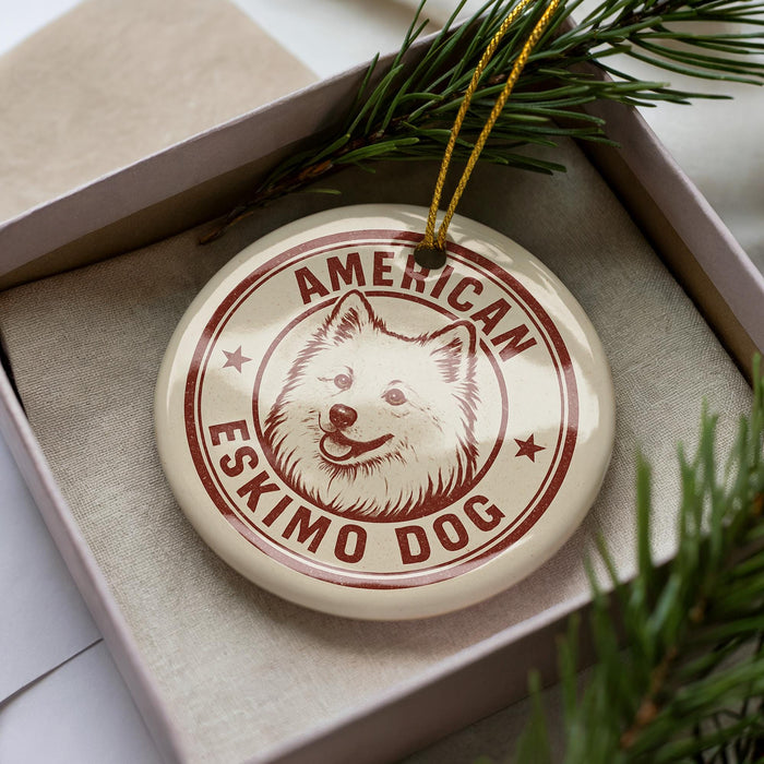 A circular ornament with a white dog's face and the text "AMERICAN ESKIMO DOG" printed on it, placed inside a box with pine branches.