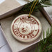 A circular ornament with a white dog's face and the text "AMERICAN ESKIMO DOG" printed on it, placed inside a box with pine branches.