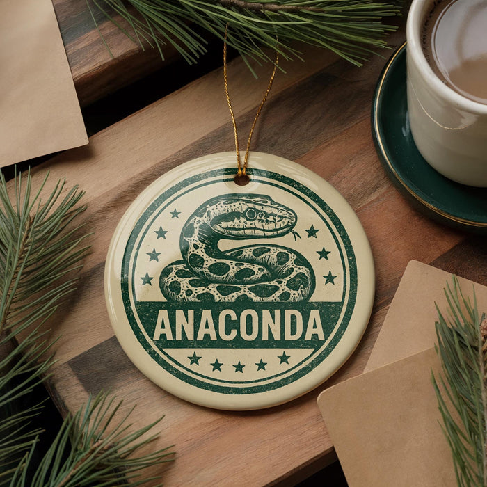A round ornament with a snake design and the word "ANACONDA" printed on it, placed on a wooden surface next to a cup of coffee and some pine branches.