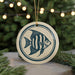 A round ceramic ornament featuring a blue and white striped fish design, hanging from a string and placed on a surface with pine branches in the background.