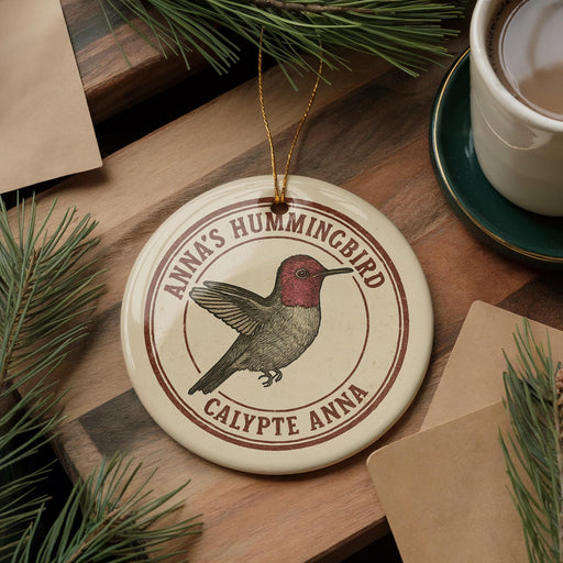 A round ornament featuring a hummingbird is displayed on a wooden surface, surrounded by pine branches and a cup of coffee.