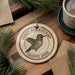 A round ornament featuring a hummingbird is displayed on a wooden surface, surrounded by pine branches and a cup of coffee.