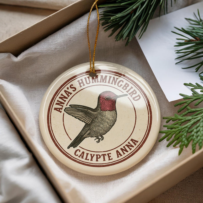A round ornament with a hummingbird image and text, placed in a box with pine branches.