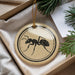 A circular ornament with a black ant illustration is displayed in a box, surrounded by pine branches and a white paper.
