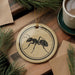 a round ornament with a black ant illustration on it, placed on a wooden surface next to a cup of coffee and some pine branches.