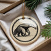 A circular ornament featuring a black and white illustration of an animal, likely a bear, is displayed in a box with pine branches.