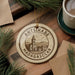 A circular ornament with the text "ANTSIRABE MADAGASCAR" and an illustration of a church, placed on a wooden surface next to a cup of coffee and pine branches.