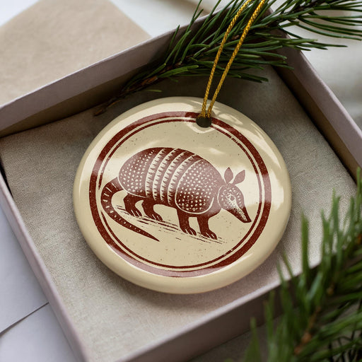 A ceramic ornament in the shape of an armadillo, with a brown and white design, is displayed in a box surrounded by pine branches.
