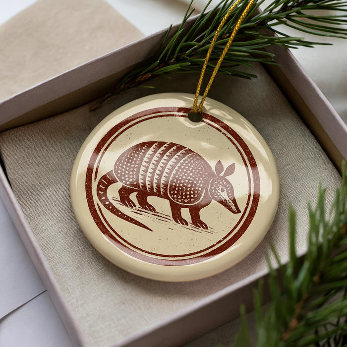 A ceramic ornament in the shape of an armadillo, with a brown and white design, is displayed in a box surrounded by pine branches.
