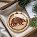 A decorative ornament featuring an armadillo is displayed in a box, surrounded by pine branches.