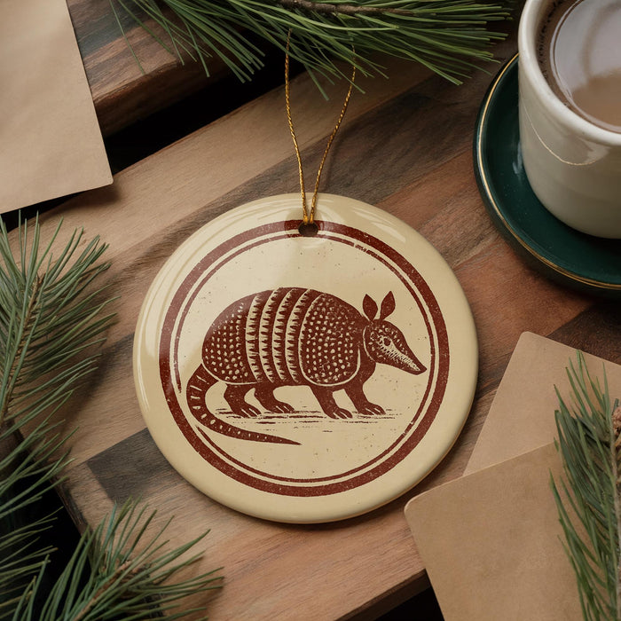 a round ornament with a brown and white design depicting an armadillo, placed on a wooden surface alongside a cup of coffee and some pine branches.