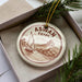 A decorative ornament in a box, featuring a sailboat and a landscape scene with mountains, all set against a brown background.