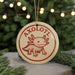 A round ornament with the word "AXOLOTL" printed on it, featuring an illustration of an axolotl, a type of salamander. The ornament is hanging from a string, and there are pine branches visible in the background.