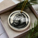 A ceramic ornament in the shape of a monkey's face is displayed in a box, surrounded by pine branches.