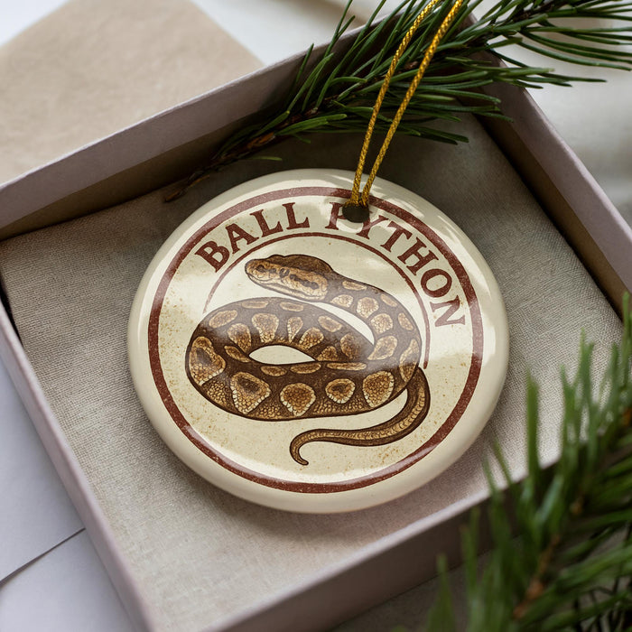A round ornament with a snake design, featuring the text "BALL PYTHON" on it, is displayed in a box with pine branches.