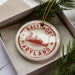 A circular ornament with the text "BALTIMORE MARYLAND" printed on it, surrounded by a decorative border. The ornament is placed inside a box, with pine branches visible in the background.