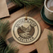 A circular ornament with the text "BALTORO GLACIER PAKISTAN" and a mountain landscape design hangs on a wooden surface, surrounded by pine branches and a cup of coffee.