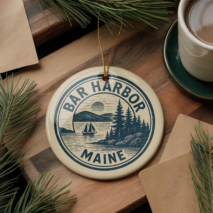 A circular ornament with the text "BAR HARBOR MAINE" and a sailing boat illustration, hanging on a wooden surface with pine branches and a cup of coffee nearby.