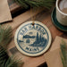 A circular ornament with the text "BAR HARBOR MAINE" and a sailing boat illustration, hanging on a wooden surface with pine branches and a cup of coffee nearby.