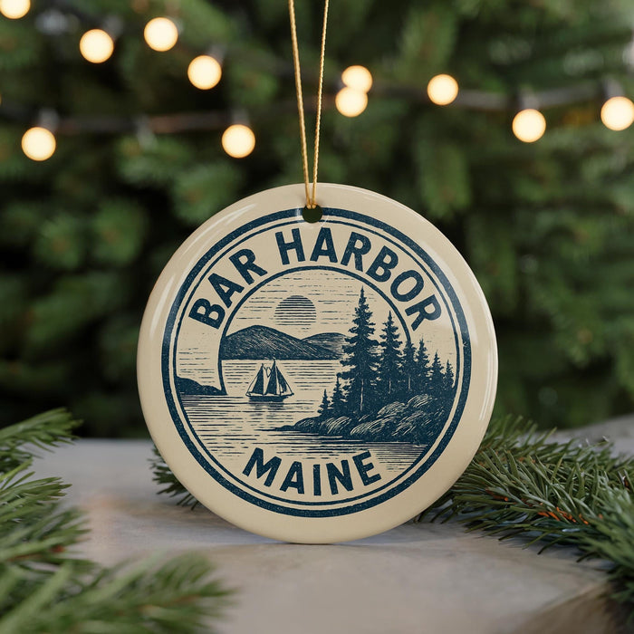 A circular ornament with the text "BAR HARBOR Maine" and a sailing boat illustration, hanging from a string against a blurred background of Christmas lights.