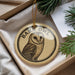 A decorative ornament featuring a barn owl is displayed in a box, surrounded by pine branches.