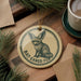 A circular ornament featuring a bat-eared fox illustration, with the text "BAT-EARED FOX" printed on it. The ornament is placed on a wooden surface, surrounded by pine branches and a cup of coffee.