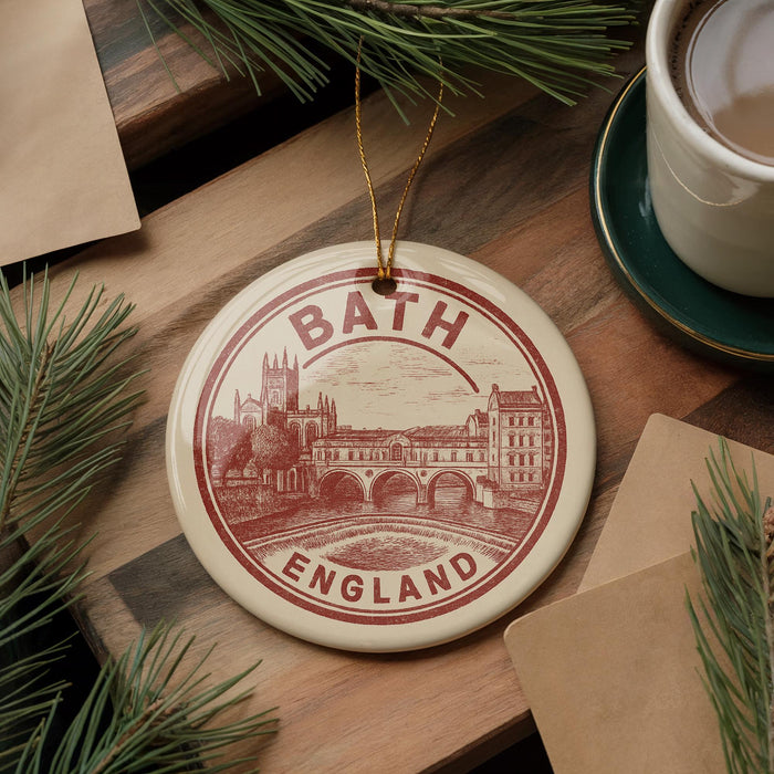 A circular ornament with the text "BATH ENGLAND" and an illustration of a cityscape is displayed on a wooden surface, surrounded by pine branches and a cup of coffee.