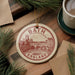 A circular ornament with the text "BATH ENGLAND" and an illustration of a cityscape is displayed on a wooden surface, surrounded by pine branches and a cup of coffee.
