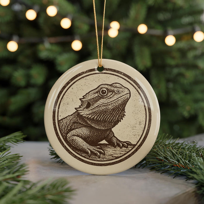 A ceramic ornament featuring a detailed illustration of a bearded dragon is displayed in front of a blurred background of Christmas lights.