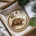 A circular ornament with a beaver illustration is displayed in a box, surrounded by pine branches.