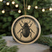 A circular ornament featuring a detailed illustration of a beetle is displayed against a blurred background of greenery and twinkling lights.