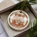 A ceramic ornament in the shape of a tiger's head, with a brown and white color scheme, is displayed in a box surrounded by pine branches.