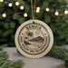 A round ornament with the text "BEQUIA SAINT VICENT AND THE GREPRADINES" printed on it, surrounded by pine branches and a blurred background of Christmas lights.