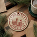 A circular ornament with the text "BISKHEK Kyrgyzstan" and an illustration of a building and a horse, hanging on a wooden surface.