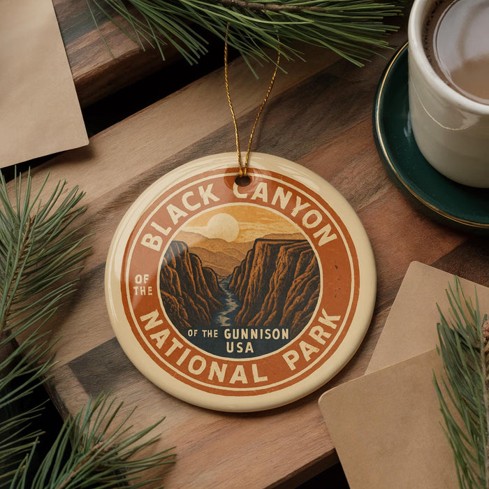 A circular ornament with the text "Black Canyon of the Gunnison National Park" and an illustration of the canyon, placed on a wooden surface alongside pine branches and a cup of coffee.