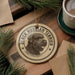 A circular ornament featuring a black Russian terrier dog's head, with the text "BLACK RUSSIAN TERRIER" printed on it, is displayed on a wooden surface alongside a cup of coffee and pine branches.
