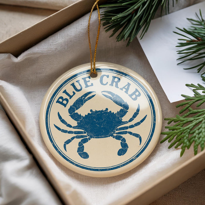 A round ornament with a blue crab design, hanging from a string, placed on a surface with pine branches and a white paper.