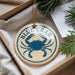 A round ornament with a blue crab design, hanging from a string, placed on a surface with pine branches and a white paper.