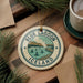 A circular ornament with the text "BLUE LAGOON ICELAND" and an illustration of a lake and mountains, hanging on a branch with pine needles.